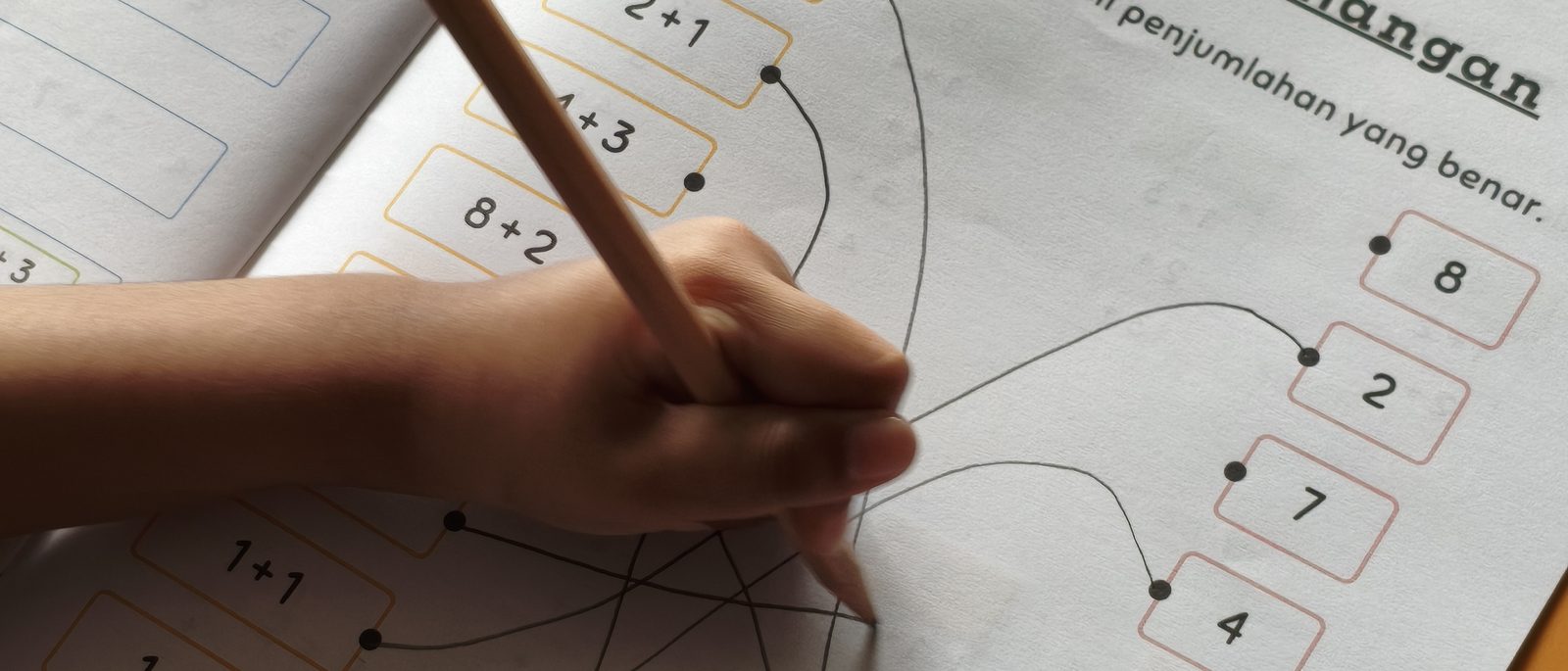 Tangan anak menarik garis jawaban dengan pensil di lembar kerja penjumlahan